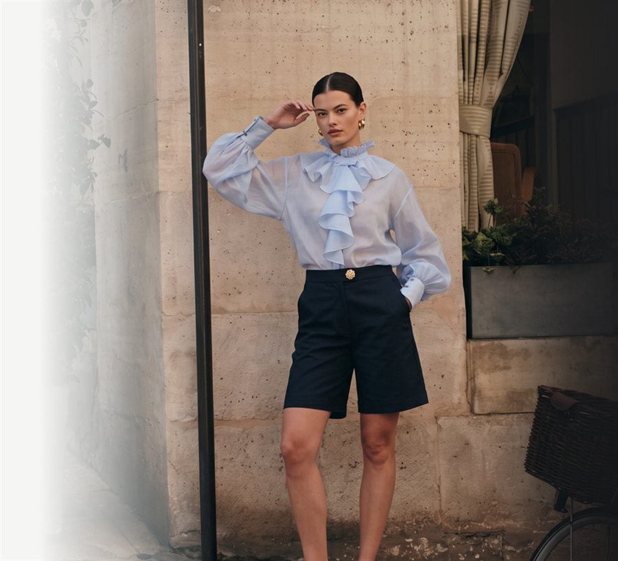 Modelo con blusa azul de volantes y bermudas azul marino junto a una pared de piedra, sesión de moda de photoeloi
